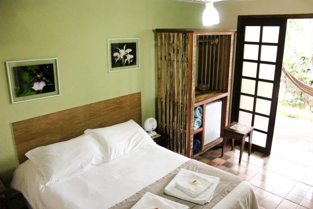Um quarto simples com uma cama de casal, guarda-roupa de madeira, mesa de cabeceira, toalhas e uma porta aberta que dá para uma rede do lado de fora, na Gamboa Eco Refúgio Pousada em Iporanga (PETAR), apra representar pousadas no interior de SP para casal.
