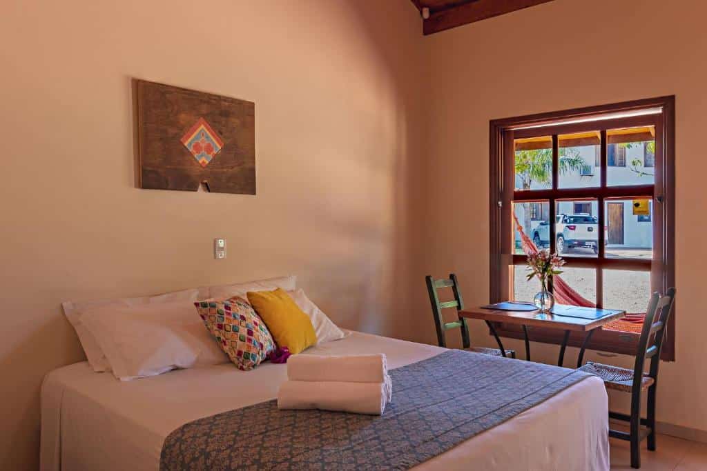 Uma cama bem arrumada com travesseiros coloridos, uma pequena mesa com cadeiras e uma janela que permite a entrada de luz natural, na Pousada Pé na Terra em Brotas, para representar pousadas no interior de SP para casal.