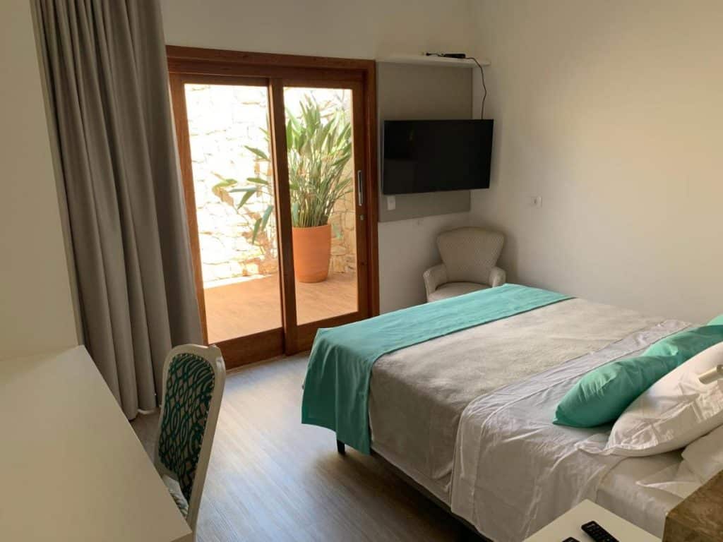 Um quarto moderno na Pousada Gardenian em São Roque com uma cama de casal, TV, cadeira, escrivaninha e portas de vidro que dão para um pátio com um vaso de plantas, para representar pousadas no interior de SP para casal.