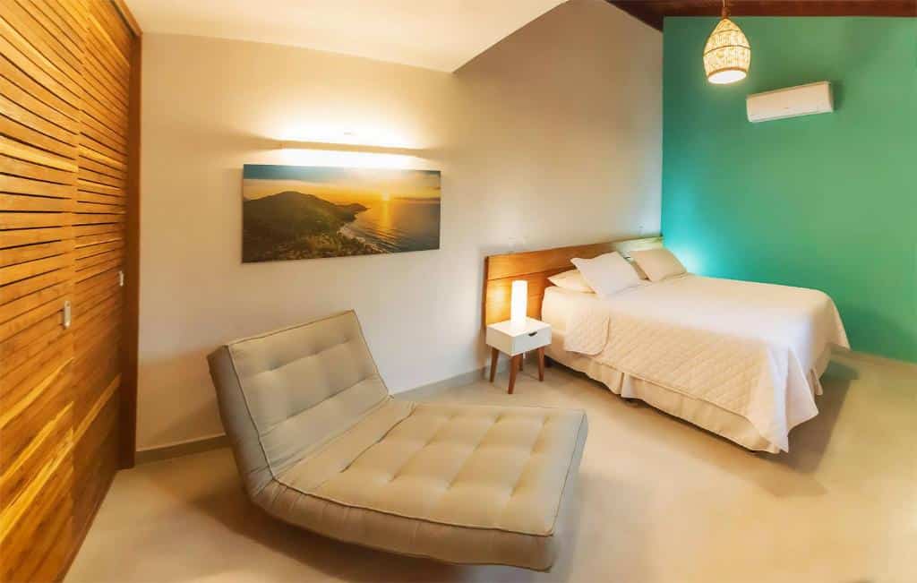 Um quarto com uma cama de casal, parede com detalhes em azul-petróleo, poltrona, mesinhas de cabeceira, arte na parede e um ar-condicionado, na Pousada Jamboo em Ubatuba, para representar pousadas no interior de SP para casal.