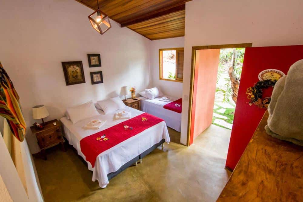 Um quarto com uma cama de casal e uma cama de solteiro, cada uma com cobertores vermelhos, uma mesa de cabeceira e uma porta aberta para um jardim, na Pousada Paineira em Bragança Paulista, para representar pousadas no interior de SP para casal.