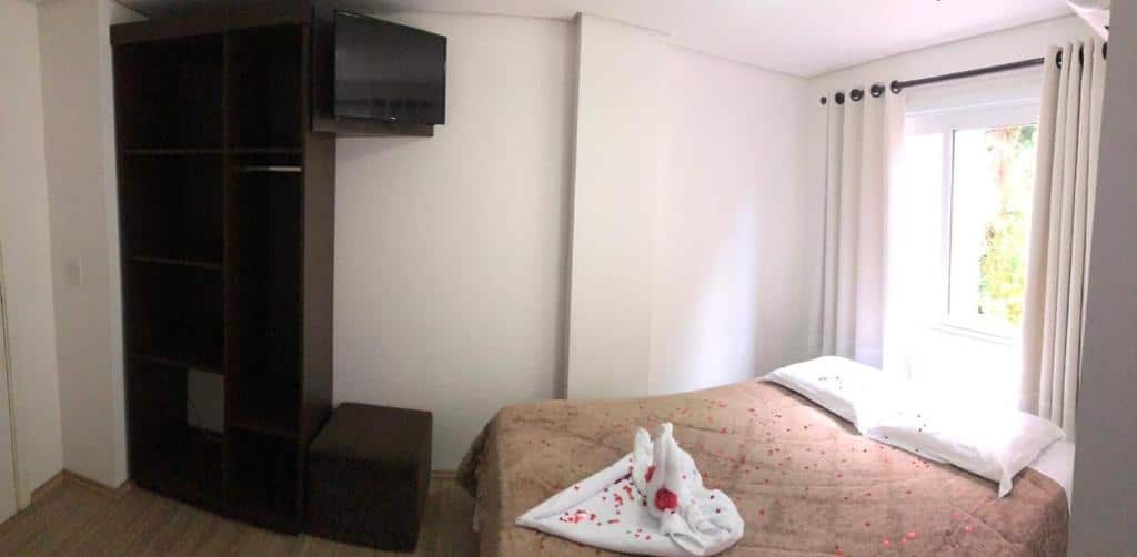 Quarto da Pousada Beija Flor Centro. Uma cama de casal do lado direito com decoração romântica e uma janela em cima. Do lado esquerdo uma televisão e um pequeno guarda-roupa.