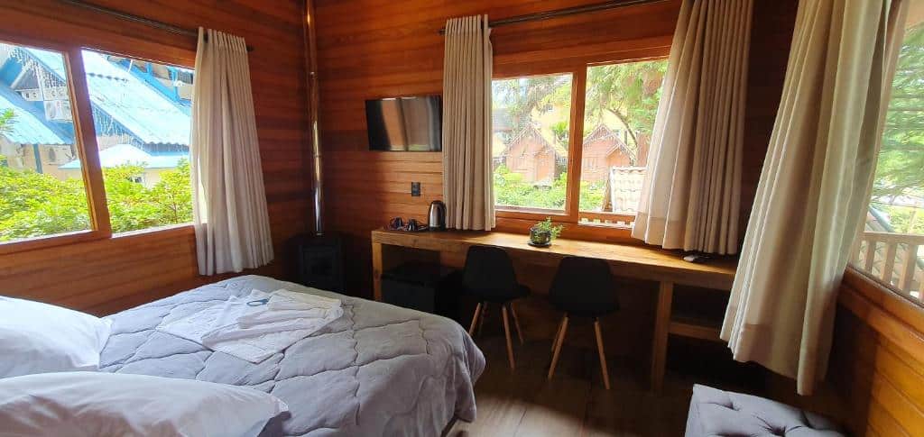 Quarto da Pousada Árvore Da Coruja. Do lado esquerdo a cama de casal, de frente a televisão, lareira e uma mesa de trabalho com duas cadeiras. As paredes com janelas de vidro, cortinas e vista das árvores. Foto para ilustrar post sobre pousadas pet friendly em Gramado.