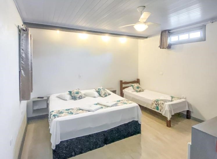 Quarto da Pousada Brisa de Gramado. No meio uma cama de casal, do lado direito uma cama de solteiro. De cada lado janelas, no teto um ventilador.