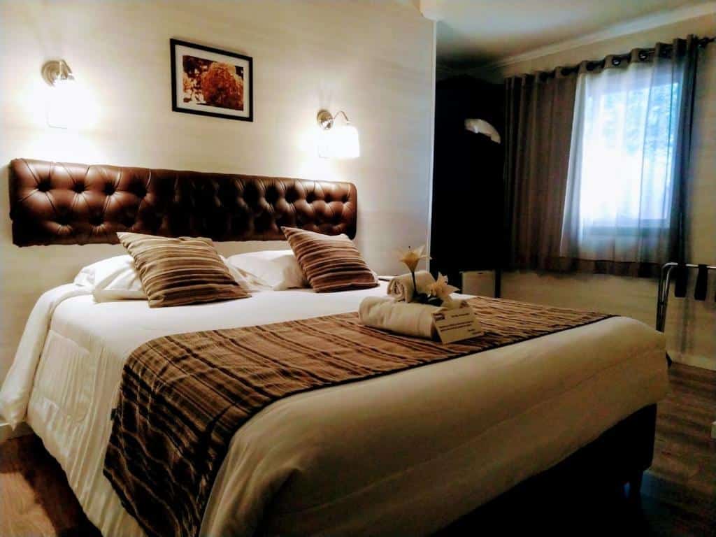 Quarto da Pousada Jasmim. Uma cama de casal com decoração em cima. No canto direito a janela com cortina fechada.