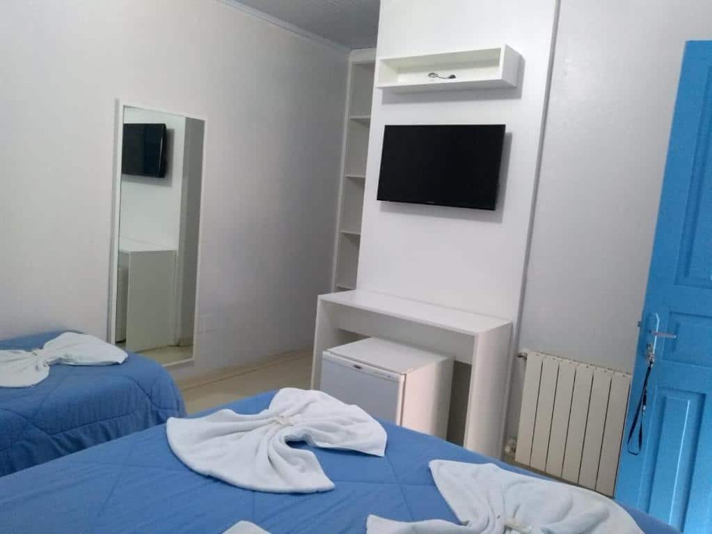 Quarto da Pousada Recanto Alpino. Uma cama de casal, do lado esquerdo uma cama de solteiro e um espelho. De frente um frigobar, uma mesa de apoio e uma televisão. Foto para ilustrar post sobre pousadas pet friendly em Gramado.