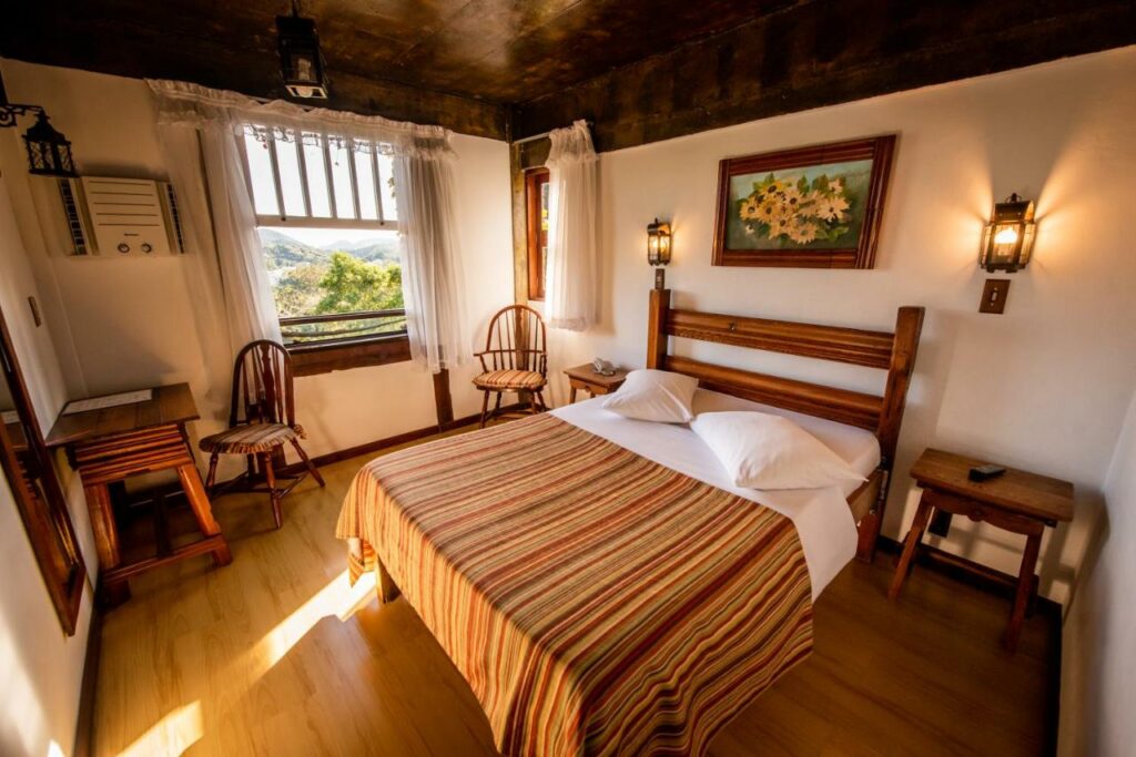 Quarto da Pousada Monte Imperial. Uma cama de casal está no meio, ao seu lado direito uma mesinha de madeira. Do lado esquerdo há uma mesa com cadeira, uma janela e outra cadeira abaixo dela. Essa é uma das pousadas pet friendly em Petrópolis.