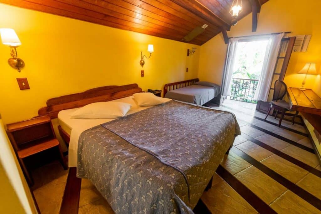 Quarto da Pousada Princesa Isabel - Visconde. Uma cama de casal e uma cama de solteiro estão lado a lado, em frente a elas uma mesa de madeira e uma cadeira. Ao fundo está a varanda.
