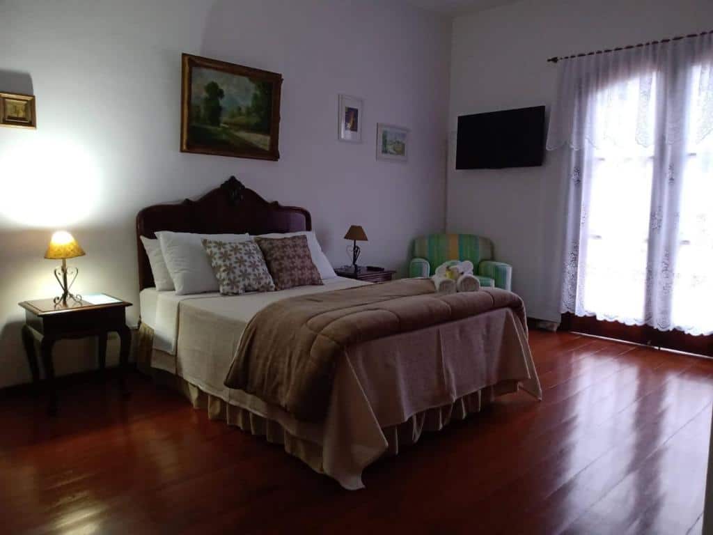Quarto da Pousada Canto da Paz. Uma cama de casal está no meio, ao seu lado direito uma poltrona, uma tv e uma janela com cortina. Ela também faz parte das pousadas pet friendly em Petrópolis.