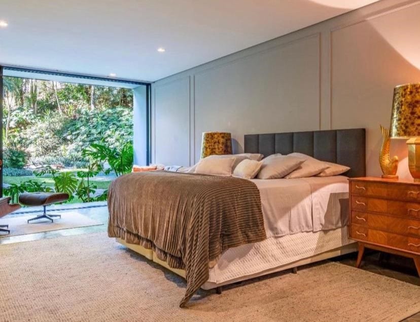 Quarto do Exclusive Oásis by Lotta De Macedo Soares. Uma cama de casal está ao lado de uma mesa de cabeceira no lado direito. Do lado esquerdo há uma parede de vidro e um banco. Essa é uma das pousadas pet friendly em Petrópolis.