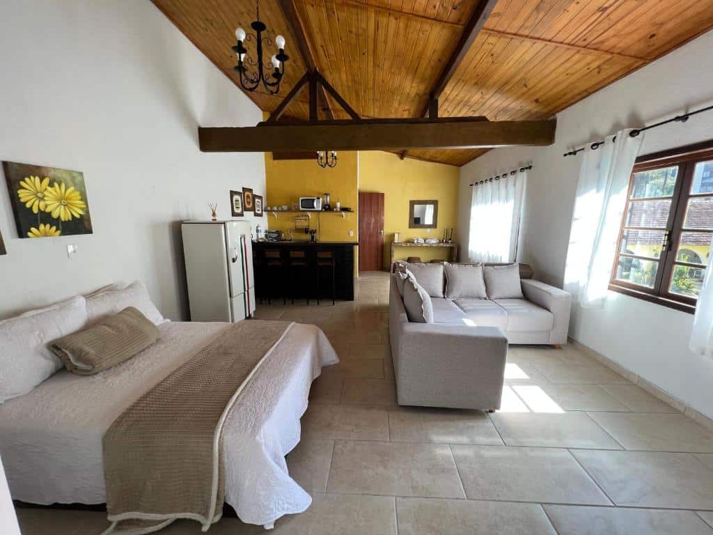 Quarto da Villa Aconchego Corrêas Itaipava. Uma cama de casal está de frente para um sofá, que está virado de frente para uma janela. Ao fundo há uma geladeira e uma cozinha compacta com balcão, mmicro-ondas e utensílios.
