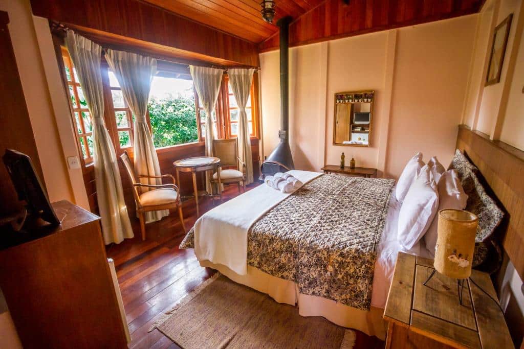Quarto da Pousada Apoema Petrópolis-rj. Uma cama de casal está de frente para uma janela, uma mesinha e duas cadeiras. Perto dela também há uma lareira. No outro canto está uma pequena tv.