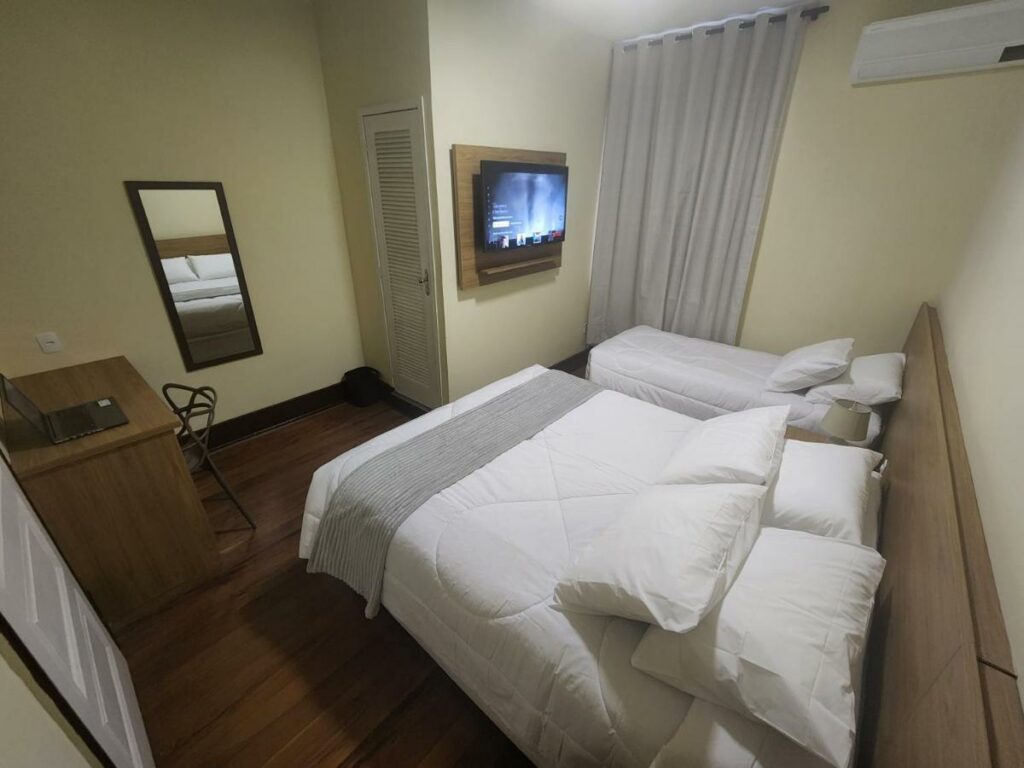 Quarto da Pousada Catedral. Uma cama de casal e uma de solteiro estão lado a lado e de frente para uma tv. Mais em frente está uma mesa de trabalho e uma cadeira, um espelho e uma porta.