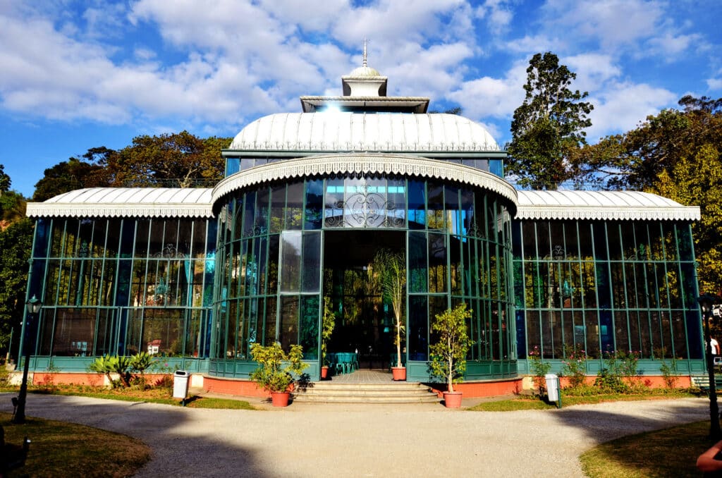 Palácio de Cristal em Petrópolis. Ele é todo transparente e azulado e possui diversas plantaspor dentro e ao redor.