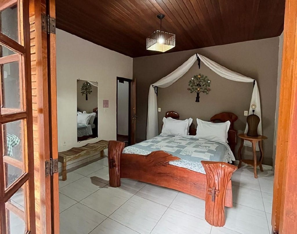 Quarto da Pousada Aconchego. Uma cama de casal está no meio, ao lado de um banco de madeira e um espelho (lado esquerdo). Ao seu lado direito há um abajur e em frente uma porta. Foto para ilustrar post de pousadas pet friendly em Pirenópolis.