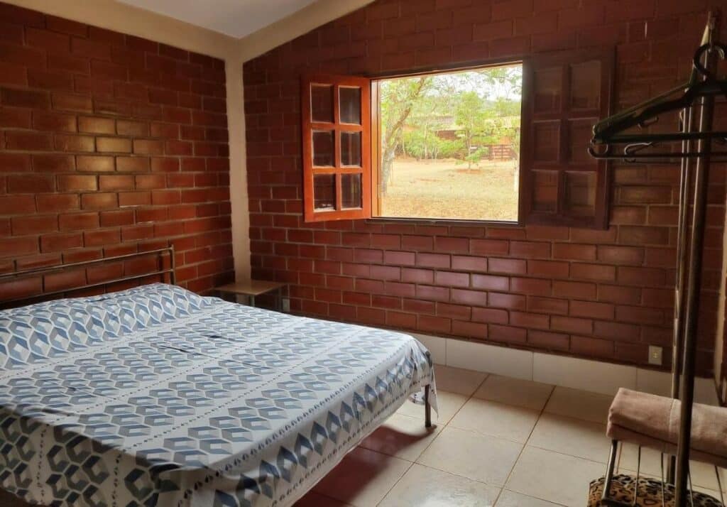 Quarto da Pousada Carmel Garden. Uma cama de casal está no meio, ao lado de uma janela. Em frente está uma arara para roupas. As paredes são de tijolos. Esta é uma das pousadas pet friendly em Pirenópolis.