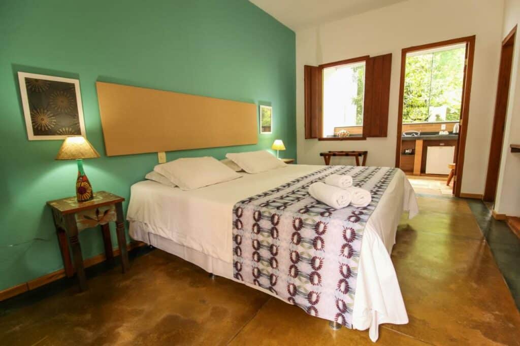 Quarto da Pousada Arvoredo. Uma cama de casal está no meio, ao seus dois lados mesinhas com abajures. Ao fundo é possível ver uma cozinha compacta na varanda.