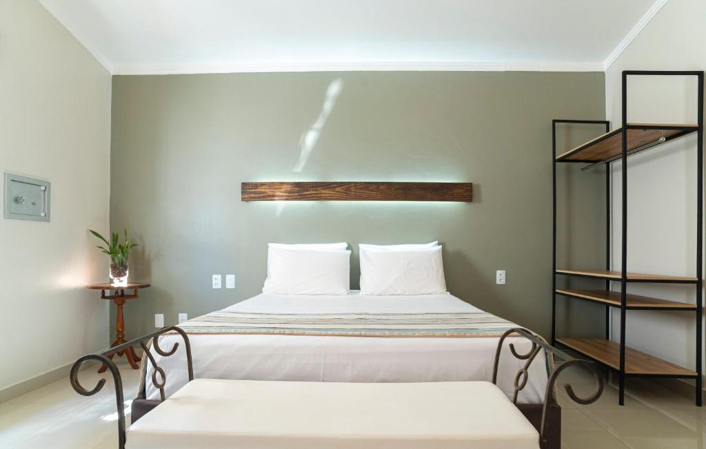 Quarto da Villaê Pousada. Uma cama de casal está no meio, ao seus pés um recamier. Ao lado direito há um armário aberto com quatro prateleiras. Do lado esquerdo há uma mesinha com abajur. Ela faz parte das pousadas pet friendly em Pirenópolis.
