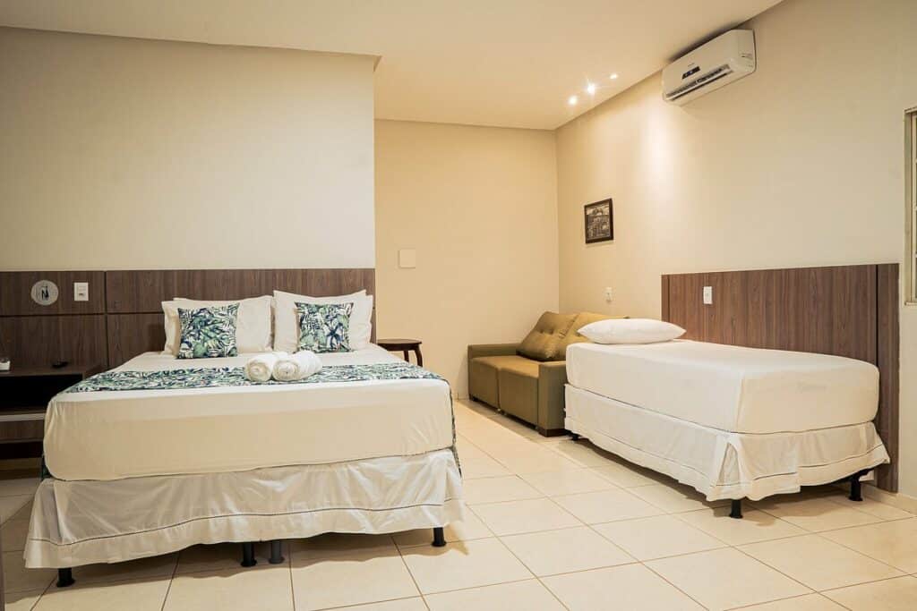 Quarto da Pousada Renascer Park. Uma cama de casal está do lado esquerdo e uma cama de solteiro está do lado direito. Atrás delas há um sofá.