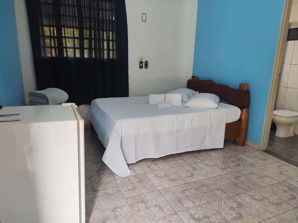 Quarto da Pousada Carvalho. Uma cama de casal está no canto esquerdo, ao lado de uma parede com janela. Perto da cama há um frigobar. A imagem faz parte da lista de pousadas pet friendly em Pirenópolis.