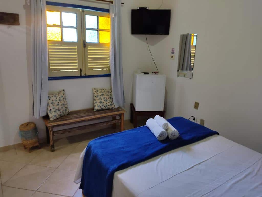 Quarto da Pousada Colonial com uma cama de casal de frente par auma parede com um banco e uma janela. Há também um frigobar e uma tv. Essa é uma ótima opção de pousadas pet friendly em Pirenópolis.