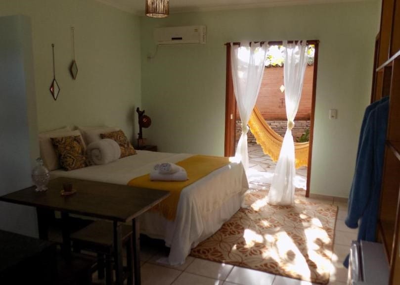 Quarto da Pousada Fênix. Uma cama de casal está de frente para um armário e ao lado de uma mesa. Ao fundo está uma porta e uma rede. A pousada Fênix é uma das pousadas pet friendly em Pirenópolis.