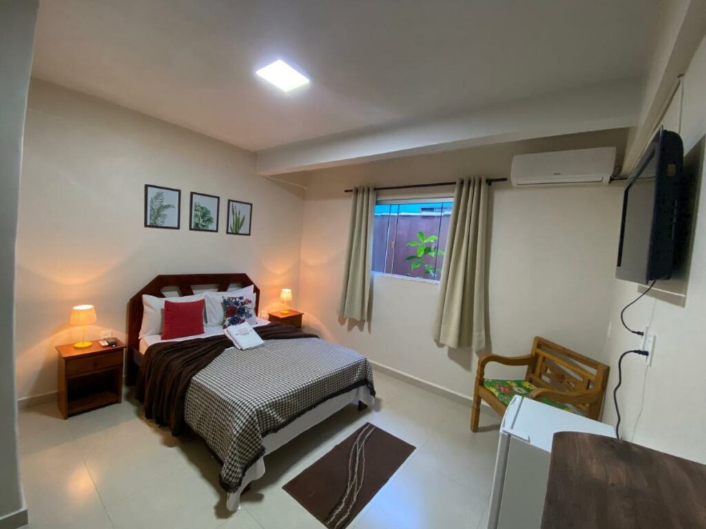 Quarto da Chico de Sá Pousada. Uma cama de casal está de frente para uma tv, um banco e um frigobar. Ao seu lado está uma janela. Dos dois lados da cama estão mesinhas com abajur. Essa é mais uma boa opção de pousadas pet friendly em Pirenópolis.