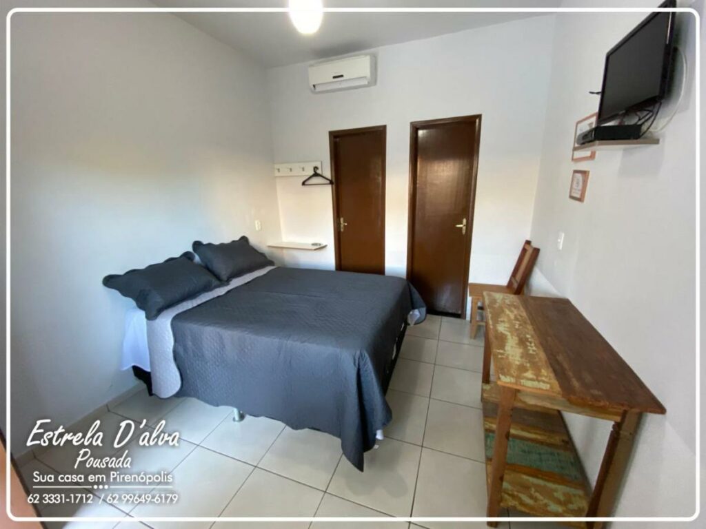 Quarto da Estrela D Alva Pousada. Uma cama de casal está de frente para uma mesa de madeira, uma cadeira e uma tv na parede. Ao seu lado estão duas portas. Ela faz parte da lista de pousadas pet friendly em Pirenópolis.