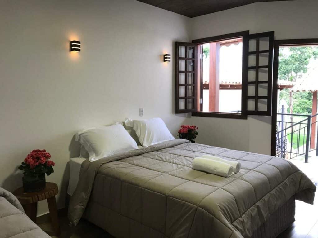 Quarto da pousada KUKA ZEN. Uma cama de casal está no meio. Do seus dois lados estão bancos com vasos de flores. Ao lado direito está uma janela.