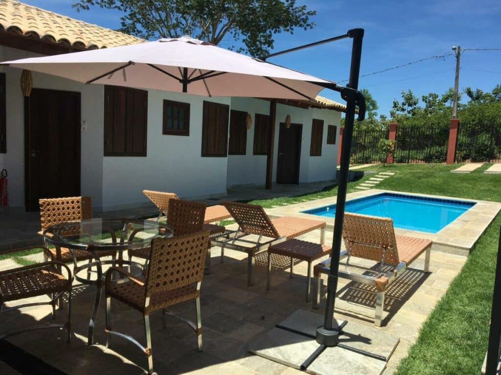 Piscina da pousada KUKA ZEN. Ela é retangular e está em uma área de pisos em meio a um gramado. Perto dela estão espreguiçadeiras, mesa e um guarda-sol.