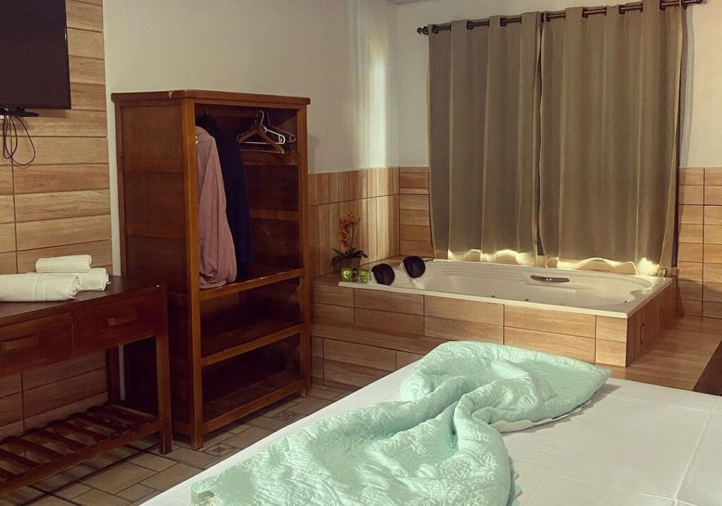 Quarto da Pousada Estrela Guia. Uma cama de casal está perto de uma banheira de hidromassagem. Ao lado da banheira há um pequeno armário para roupas. Em frente a cama está uma tv.