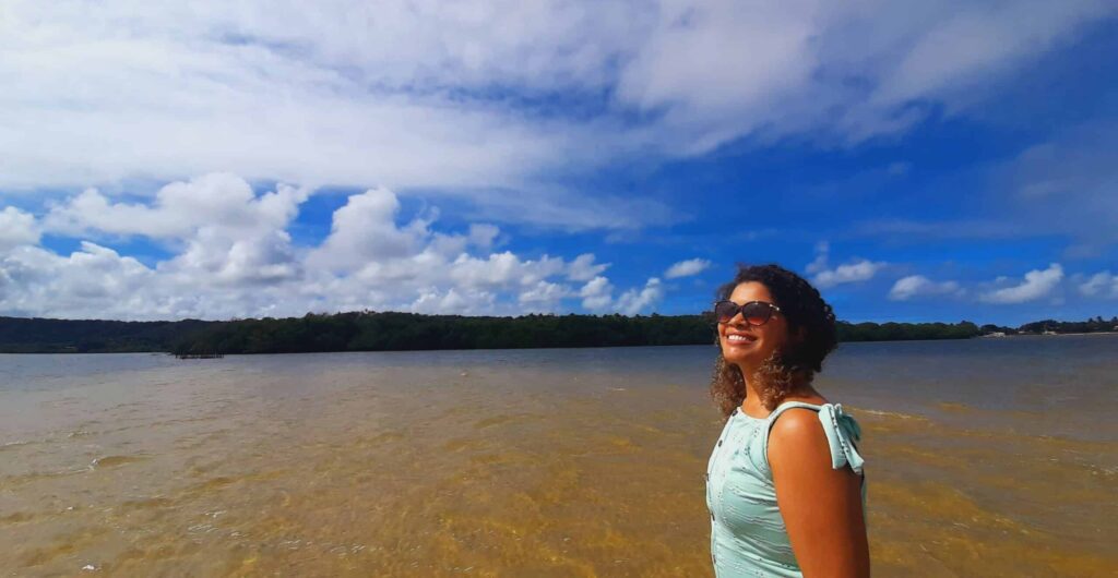 Mulher olhando para o lado, com o cabelo cacheado solo, com a lagoa do Roteiro de fundo e árvores ao fundo.
