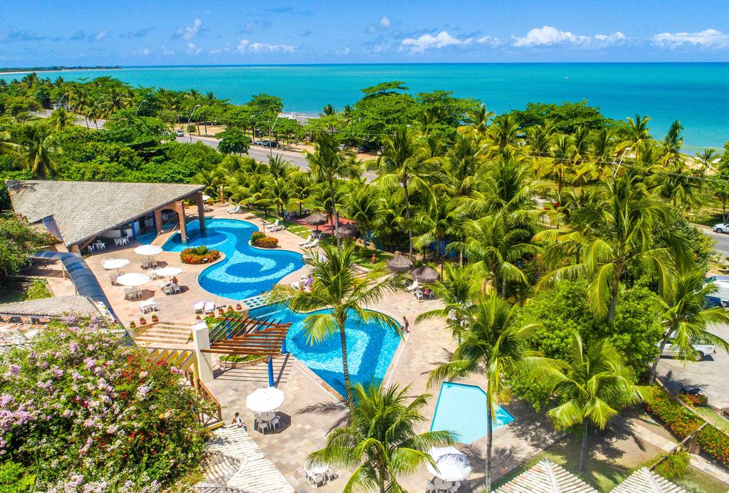 Vista aérea da parte externa com piscinas e coqueiros no Portobello Praia resort em Porto Seguro, uma estrada e, mais atrás, o mar turquesa do destino