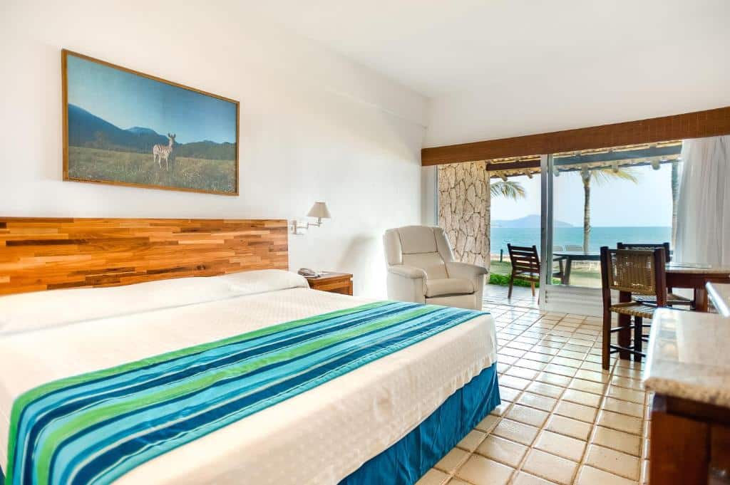 Quarto de hotel com uma cama grande, poltrona, escrivaninha e portas de pátio com vista para o oceano e as palmeiras do lado de fora.