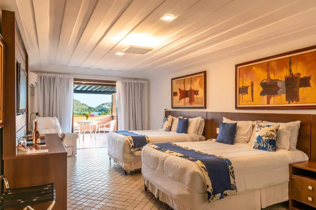 Quarto de hotel com duas camas, um sofá, mesa e cadeiras, quadros grandes e uma porta do pátio que dá para uma vista externa.