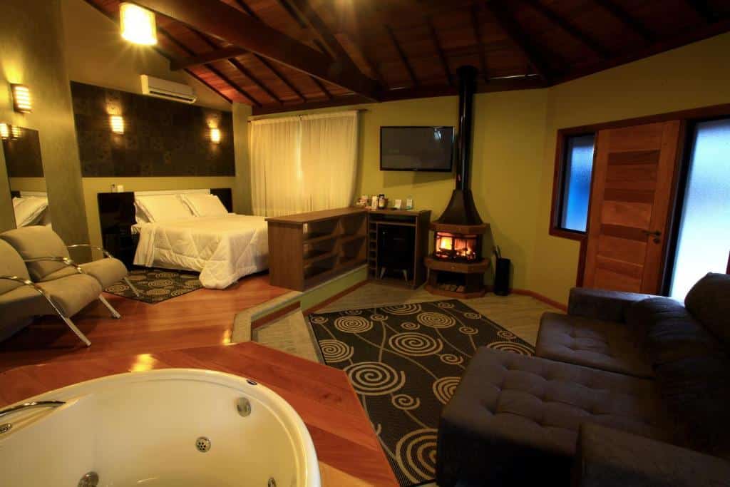 Um quarto de hotel aconchegante com uma cama, sofá, lareira redonda, TV, poltronas e uma jacuzzi em primeiro plano.