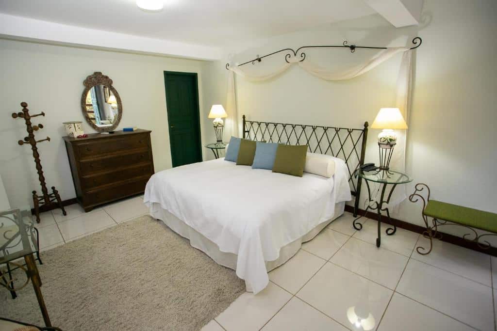 Um quarto com uma cama grande, duas mesas laterais com abajures, uma cômoda com espelho e um cabide sobre um piso de azulejos.
