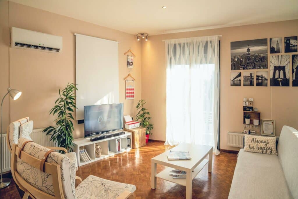 Interior de uma sala de estar com tacos no chão, uma poltrona à direita e, à esquerda, um sof´. Há uma mesa de centro e um balcão com TV e vasos de flotes nos lados.