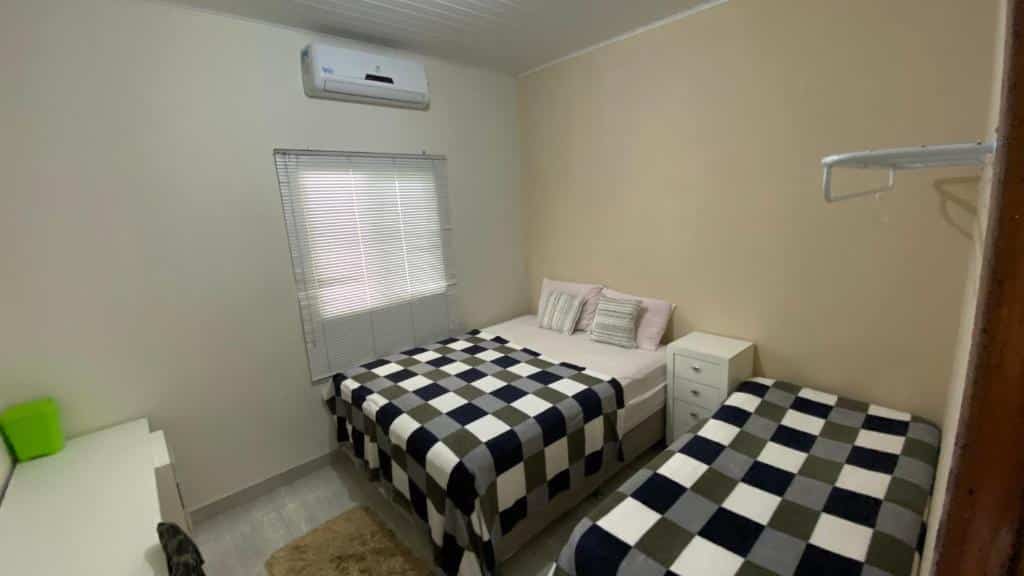 Quarto da Casa Aconchegante com Piscina. Do lado direito uma cama de solteiro e uma de casal, entre elas uma cômoda. De frente uma escrivaninha. No fundo uma janela e em cima um ar-condicionado.