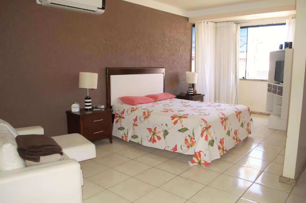 Quarto da Casa Atalaia. Do lado esquerdo um sofá, no meio uma cama com uma cômoda de cada lado e um abajur. Do lado direito uma raque com televisão e uma janela de vidro com cortina. Foto para ilustrar post sobre airbnb em Aracaju.