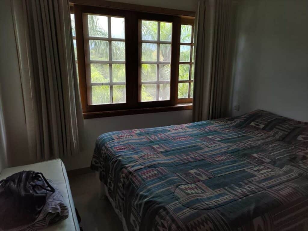 Quarto do Lindo Loft na Praia de Barra do Una à beira do Rio, um dos airbnb em Barra do Una. Uma cama de casal está encostada na parede direita e encara um apoio com mala. Ao lado esquerdo há uma janela com cortinas.