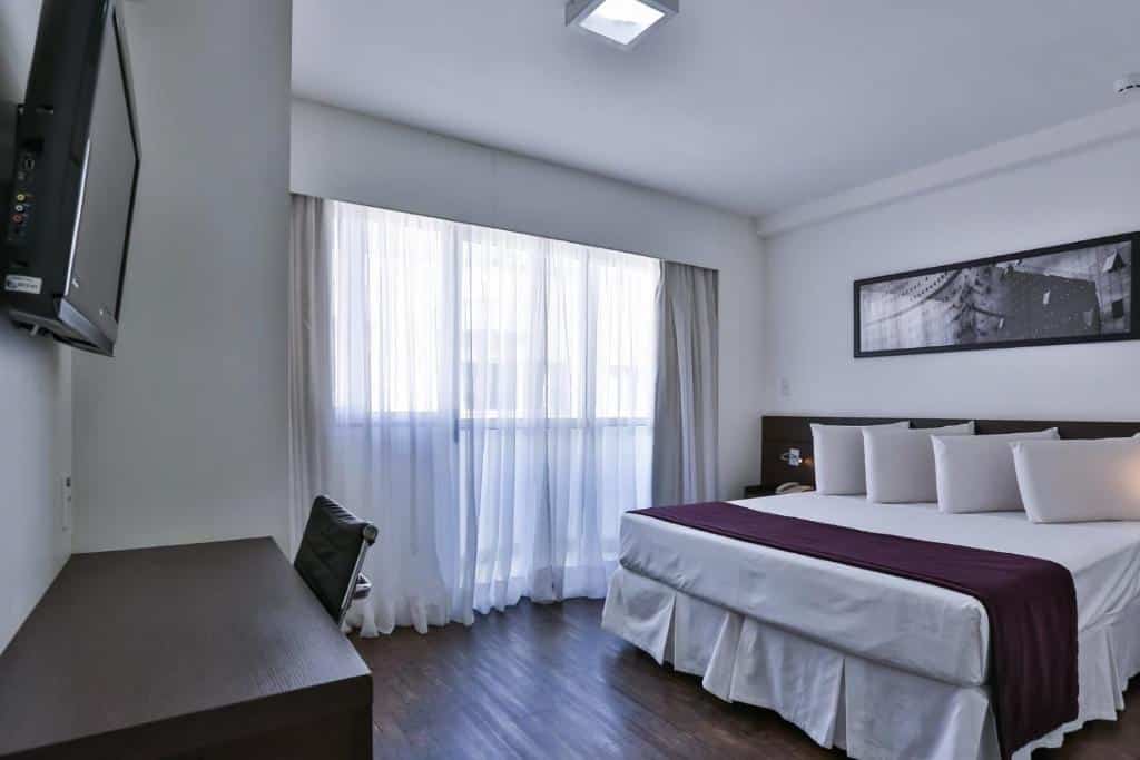Foto do quarto no Flat com Varanda-Pampulha. Há uma cama box de casal na direita da foto e um quadro atrás dela na parede. Na sua esquerda, há uma janela com cortina. No canto inferior direito, tem uma mesa com cadeira e uma TV fixa na parede. Representa o post de airbnb em Belo Horizonte.