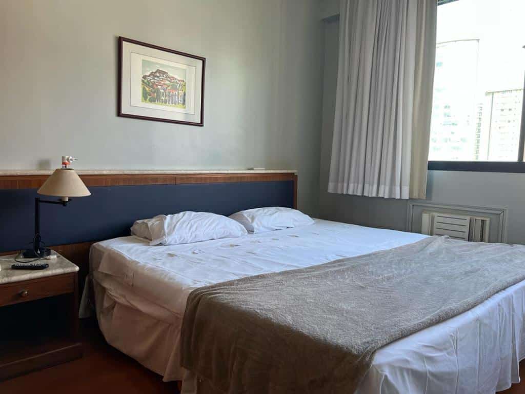 Quarto no Apart Hotel Champagnat. Há uma cama de casal no meio da foto, e uma mesa de cabeceira na esquerda com abajur. Na direita,, está a janela com cortina. Representa o post de airbnb em Belo Horizonte.