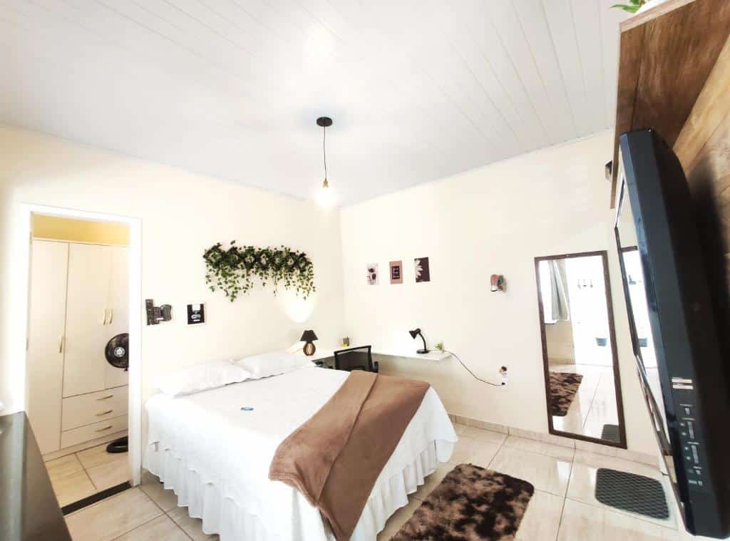 Quarto com cama box de casal da Kitnet Sol que representa o post de airbnb em Belo Horizonte. A cama está no centro, encostada na parede. Do seu lado direito há uma escrivaninha com abajur e cadeira, e do esquerdo, um espelho. No canto inferior direito da foto, de frente para a cama, há um painel com televisão. 