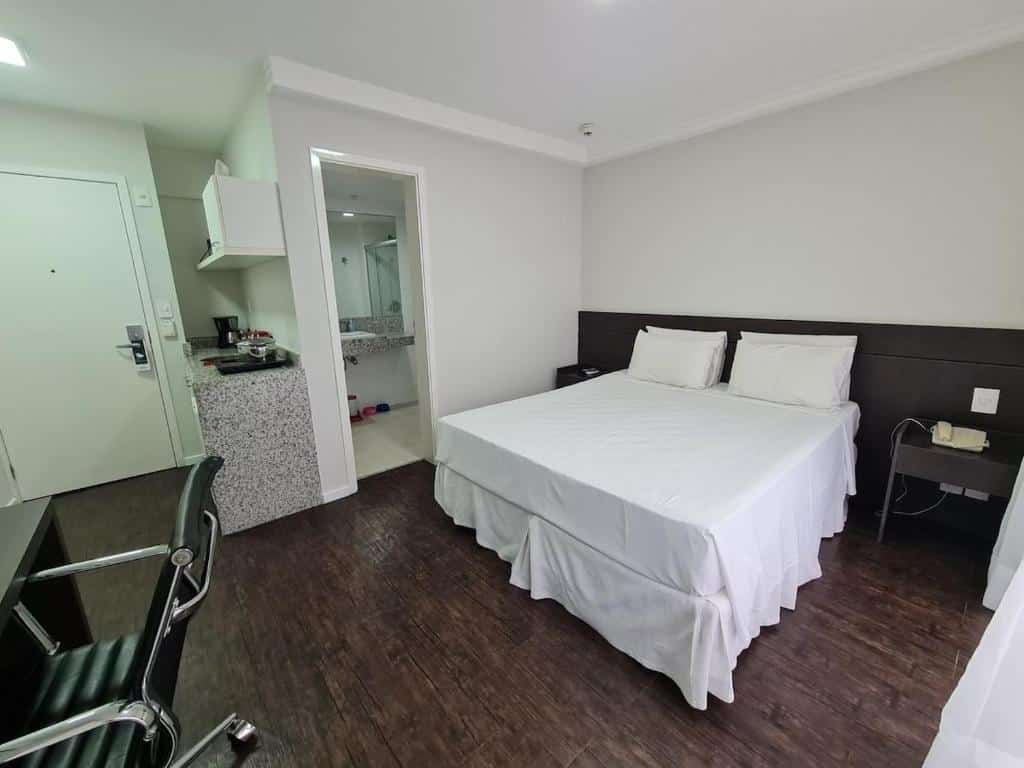 Quarto do Wafeh Flats em San Diego Pampulha. A cama é box de casal e está encostada na parede na direita, e atrás dela há uma cabeceira preta. Do lado esquerdo da cama, há uma porta para o banheiro. No canto inferior esquerdo há uma escrivaninha com cadeira. A porta está ano fundo à esquerda, e á uma mini cozinha do lado. Representa o post de airbnb em Belo Horizonte.