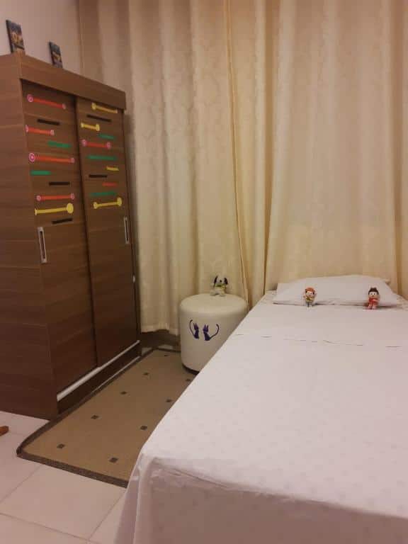 Um quarto em Pertim de Tudo com uma cama de solteira à direita e armário à esquerda. Atrás dos móveis, na parede, há uma janela com cortina.