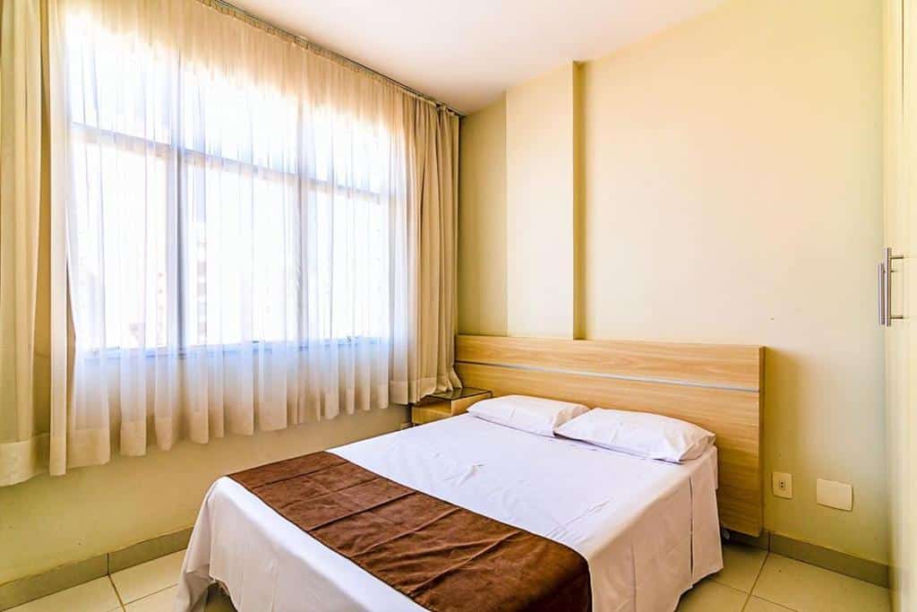 Quarto iluminado e minimalista no Villa Neves Residence. A cama de casal está no centro da foto. A cabeceira atrás dela é de madeira. Na sua esquerda há uma janela e na direita, um guarda-roupa. Representa o post de airbnb em Belo Horizonte.