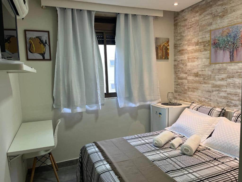 Quarto no Studios Coração Eucarístico. No canto inferior direito vemos uma cama de casal com toalhas em cima, e tem um frigobar no seu lado à esquerda. Do lado do frigobar, está a janela. No canto esquerdo, tem uma mesa de escrivaninha pequena com cadeira, e também uma TV fixa na parede. Representa o post de airbnb em Belo Horizonte.