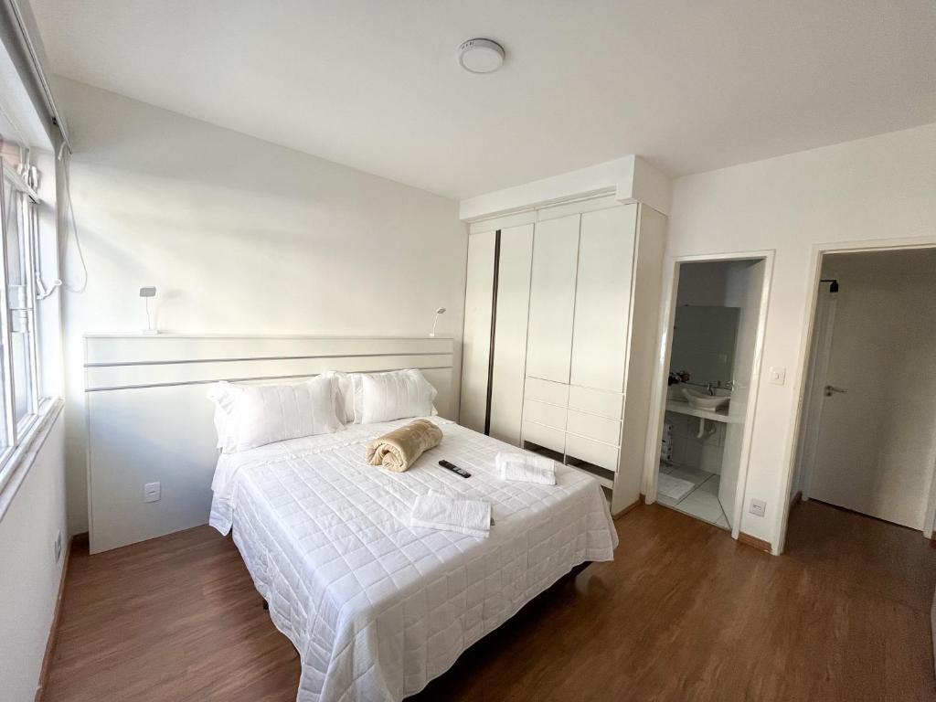 Quarto espaçoso no Apartamento em Belo Horizonte. As paredes são brancas e o piso é de madeira. Há uma cama box de casal no centro, com toalhas de banho em cima. Atrás dela há uma cabeceira com luminárias e tomada. Um guarda-roupa está na direita da cama, ao lado da porta para o banheiro do quarto. Na esquerda está a janela.