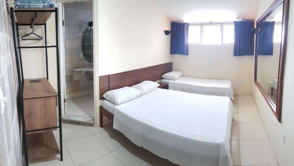 Foto no quarto na Sleep Suítes. Tem uma cama box de casal e uma de solteiro do seu lado direito, no fundo do quarto, do lado da janela. Há um espelho na frente das duas. Na esquerda, há uma arara para roupas e porta para o banheiro. Representa o post de airbnb em Belo Horizonte.